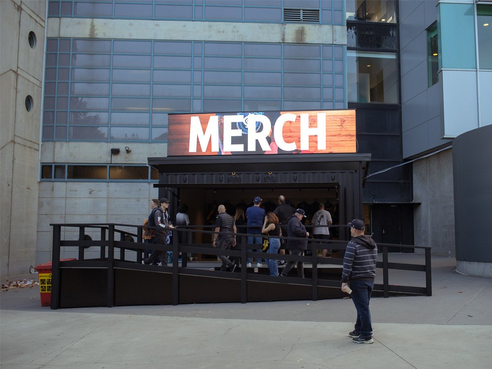 An outdoor LED billboard on a merch stall reading MERCH