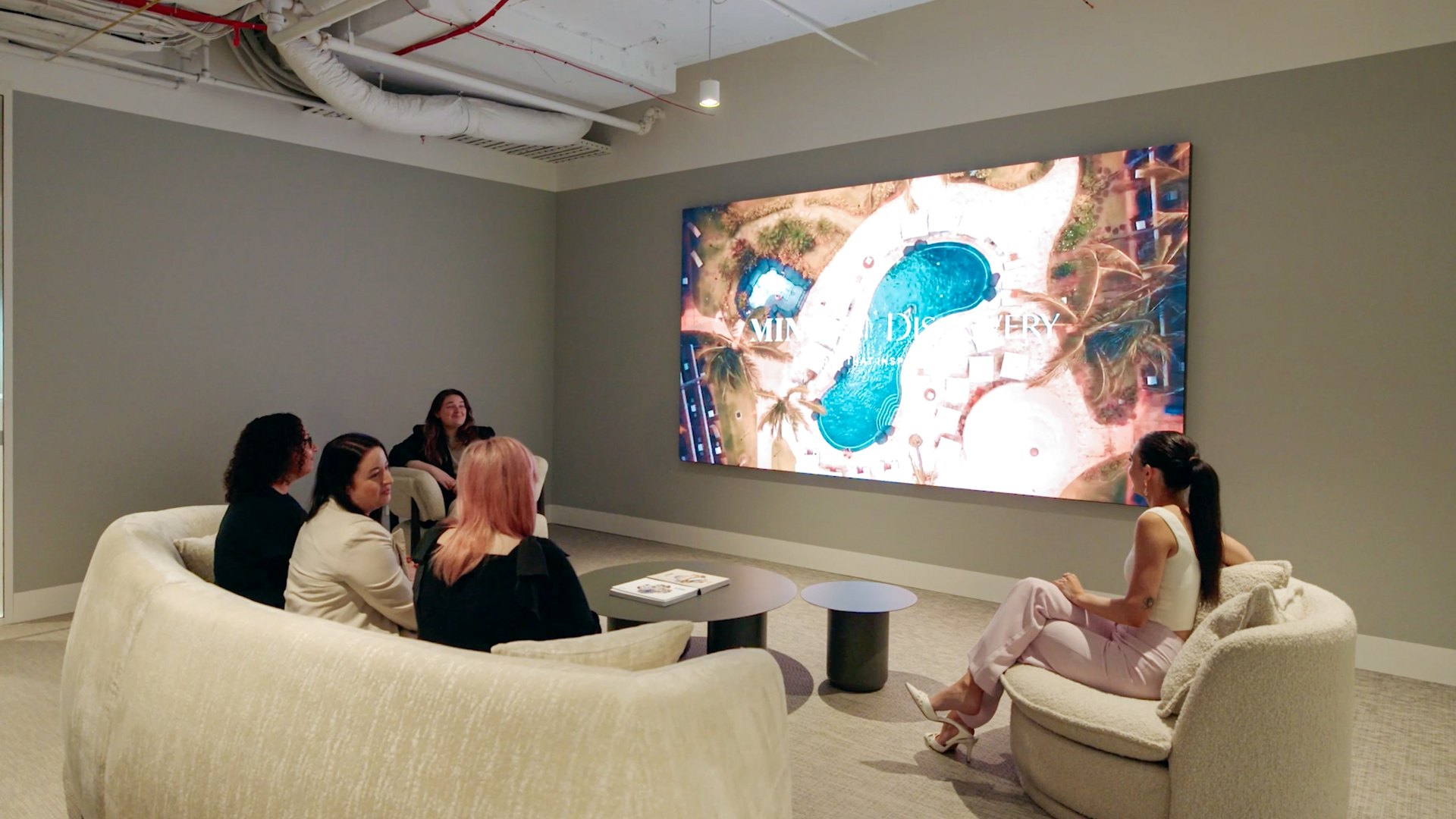 Guests in a hotel lounge watching a large LED video wall that shows an aerial view of a resort pool